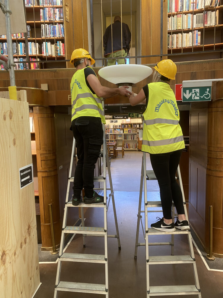 Två personer i reflexväst reparerar lampa i Stockholms stadsbibliotek 